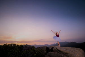 A ballet dancer wearing a leotard and tutu, standing on a rock, doing a ballet pose with a sunset background